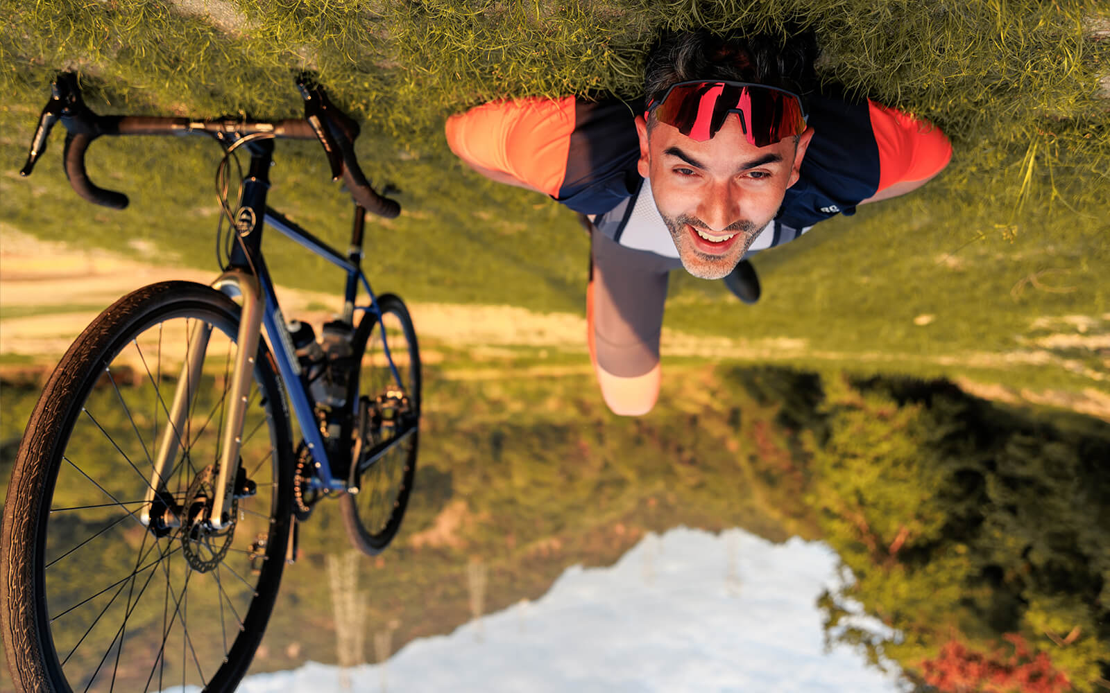 Smiling male cyclist resting on the grass with his bike after a scenic ride, reconnecting with nature – ROCKBROS Road to Sky
